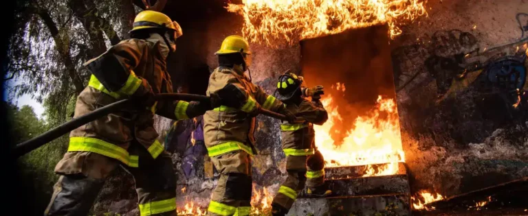 Three fire fighters carrying a hose to the mouth of a burning building.