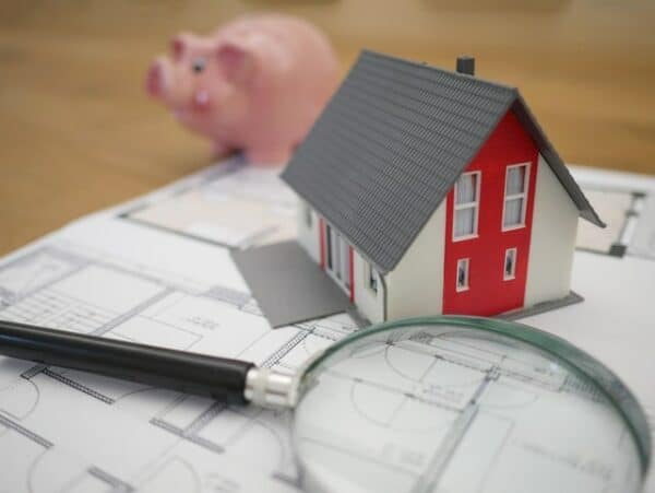 House plans, a magnifying glass, a small house model, and a piggy bank are laid out on a table.